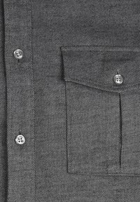 Chemise en tissu gris avec des rayures diagonales blanches, dotée d'une poche à rabat boutonnée et de deux boutons visibles sur le devant.