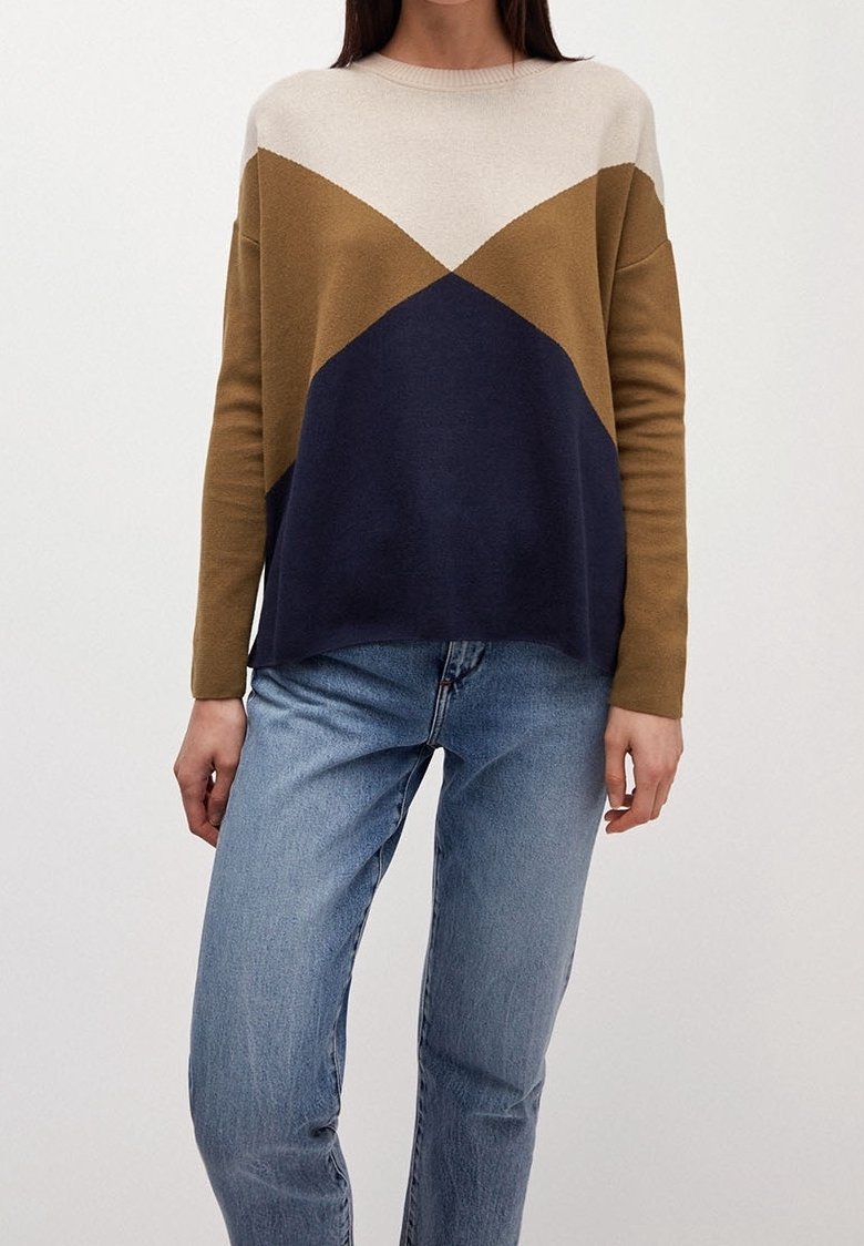 Femme portant un pull à blocs de couleurs crème, marron et bleu marine avec un jean bleu, debout devant un fond clair uni.