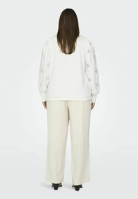 Pullover bianco in cotone con dettagli ricamati sulle maniche, abbinato a pantaloni beige chiaro a gamba larga. Tessuto morbido, vestibilità casual e silhouette ampia.