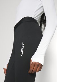 Fekete, magas derekú leggings sima textúrával. A combon fehér "TERREX" logó található, és egy fehér, hosszúujjú felsővel van párosítva.