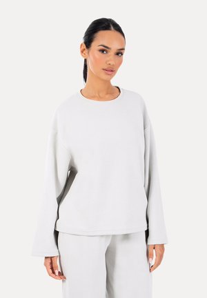 Donna con capelli scuri raccolti in una coda bassa, indossa una maglia larga a maniche lunghe di colore grigio chiaro e pantaloni abbinati, in piedi davanti a uno sfondo bianco.