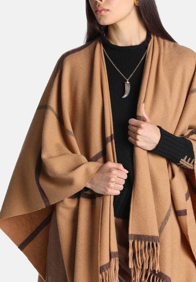 Femme portant un pull noir et un châle à carreaux marron, tenant les bords du châle des deux mains, portant un collier avec un pendentif en forme de corne.