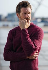Weinroter, gerippter Pullover mit langen Ärmeln, der eine figurbetonte Passform und strukturierte Stoffqualität aufweist. Das Modell legt eine Hand an das Kinn.