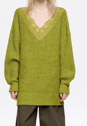 Personne portant un pull en maille vert oversize avec un col en V bordé de dentelle et un pantalon marron ample, debout sur fond blanc.