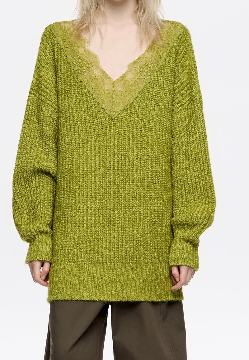 Personne portant un pull en maille vert oversize avec un col en V bordé de dentelle et un pantalon marron ample, debout sur fond blanc.