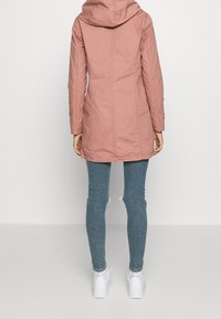 Růžová parka s kapucí a rovný střih. Obsahuje prošívané detaily a hladkou texturu, doplněná o modré skinny džíny a bílé tenisky.