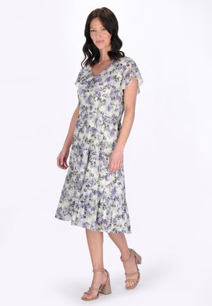 Kobieta stojąca w sukience do kolan w kwiatowy wzór z krótkimi rozkloszowanymi rękawami i beżowymi sandałami na słupku na białym tle.