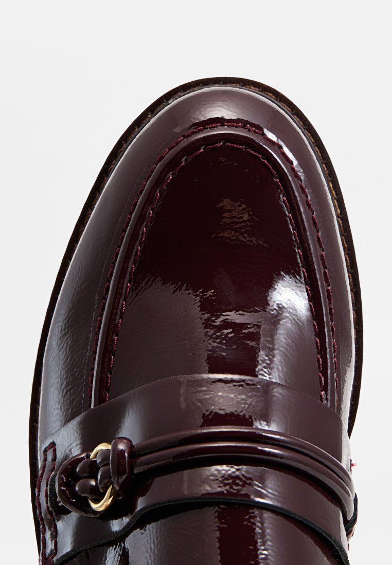 Mocassins en cuir verni bourgogne présentant un design élégant, une texture lisse et une sangle décorative avec un accent doré.