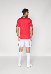 Camisola atlética vermelha com detalhes em preto, combinada com calções brancos com riscas laterais pretas, e meias brancas até ao joelho com riscas pretas.