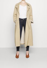 Trench coat largo beige sobre una blusa blanca, jeans oscuros y zapatos de tacón marrones. El abrigo presenta solapas anchas y mangas enrolladas.
