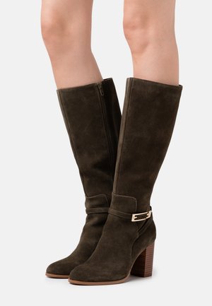 Bottes montantes en daim marron avec talons carrés, fermeture éclair latérale et sangle décorative avec boucle dorée portées sur des jambes nues.
