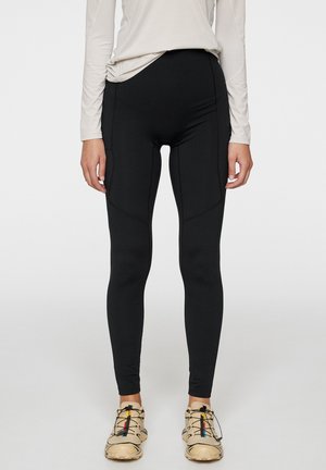 Zwarte legging met een hoge taille, een gladde textuur en naaddetails, gecombineerd met beige sportschoenen met gekleurde accenten.