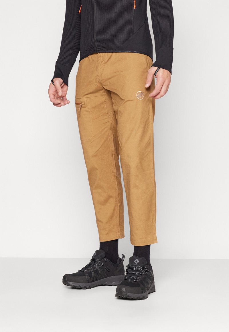 Brown cargo pants with zip pockets, tapered fit, and logo detail. Black athletic shoes with textured upper and laces, worn with dark socks.