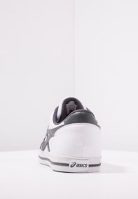 Valkoinen urheilujalkine, jossa on harmaat yksityiskohdat, synteettisestä nahasta valmistettu päällinen, pehmustettu nilkkapanta ja musta pohja. "Asics"-logo näkyvissä kantapäässä.