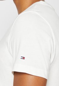 Weißes Baumwoll-T-Shirt mit Rundhalsausschnitt und kurzen Ärmeln. Mit einem kleinen gestickten Logo in Rot, Weiß und Blau auf dem linken Ärmel.