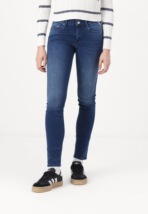 Personne portant un jean slim bleu, des chaussettes blanches montantes, des baskets Adidas noires avec des rayures blanches, et un pull blanc avec des rayures horizontales grises.