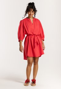 Rotes knielanges Kleid mit einem knopfbaren Kragen, langen Puffärmeln und einem taillierten Gürtel. Trägt schwarze Sandalen mit Blumenakzenten.
