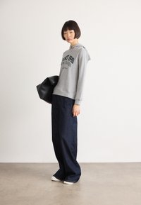Femme portant un sweat à capuche gris avec du texte, un jean bleu foncé large, des chaussures blanches, tenant un grand sac noir, se tenant devant un mur uni.