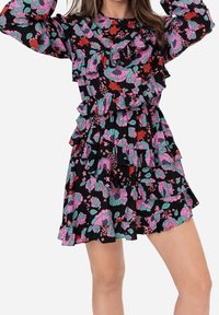 Femme portant une robe noire à fleurs roses, rouges et turquoise, avec des volants superposés et de longues manches bouffantes.