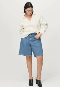 Pulover alb cu dungi, decolteu în V; pantaloni scurți din denim albastru deschis; saboți negri. Modelul stă în picioare, cu o mână pe șold, privind în jos.