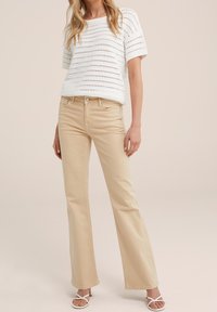 Vit stickad topp med horisontellt utskuret mönster, tillsammans med ljusbeiga utsvängda jeans och vita sandaler med remmar. Släta, lätta texturer.