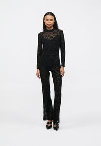 Salopetă neagră din dantelă cu mâneci lungi, model floral, bust ajustat, pantaloni evazați și guler înalt. Asortată cu pantofi negri cu vârf ascuțit.