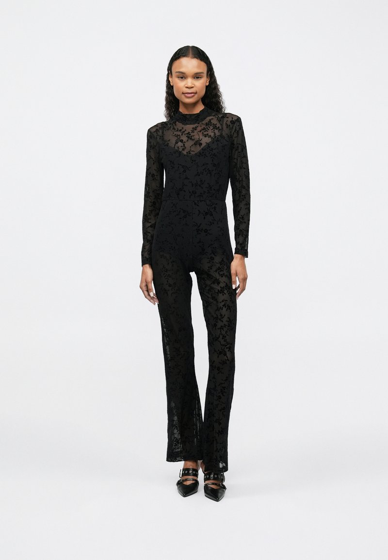 Salopetă neagră din dantelă cu mâneci lungi, model floral, bust ajustat, pantaloni evazați și guler înalt. Asortată cu pantofi negri cu vârf ascuțit.