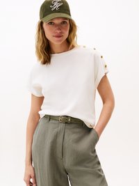 White short-sleeve top with gold button accents on shoulders, paired with light green, textured high-waisted trousers and matching belt.