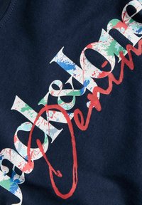 Έγχρωμα γράμματα σε μπλοκ με πιτσιλιές που σχηματίζουν τη λέξη «Brooklyn» σε σκούρο ύφασμα με επικάλυψη κόκκινου καλλιγραφικού γραψίματος και μεγάλο κόκκινο οβάλ που περικλείει μέρος του κειμένου.