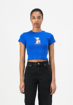 T-shirt coupé bleu en coton avec des manches courtes, arborant un imprimé graphique qui dit "OH HONEY!" en rose et blanc. Associé à un jean noir.