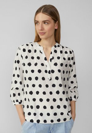 Blouse blanche à pois noirs, avec un décolleté en V et des manches trois-quarts, fabriquée à partir d'un tissu léger et texturé.