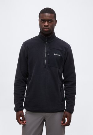 Homme portant un pull polaire noir Columbia avec une poche poitrine zippée, debout devant un fond gris uni.