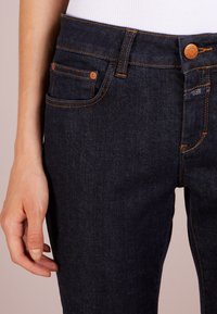 Tmavě modré džíny s oranžovým prošíváním, design s pěti kapsami a zapínáním na knoflík. Mají blízko k tělu se hodící siluetu a hladkou texturu.