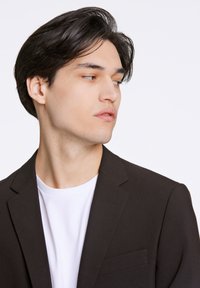 Zwarte blazer met een notched revers, een enkele borstzak, gedragen over een wit T-shirt. Soepele stof en een getailleerde pasvorm, het model kijkt opzij.