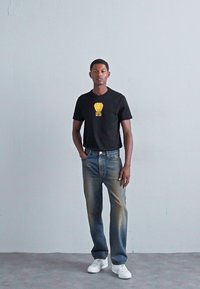 Mann, der an einer schlichten Wand steht, trägt ein schwarzes T-Shirt mit einem goldenen Löwenkopf-Design, eine blaue Jeans und weiße Sneakers, die Hände in den Taschen.