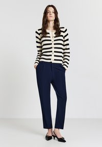 Striped black and cream cardigan with gold buttons, paired with navy tapered trousers and black slingback heels. Smooth texture, fitted design.