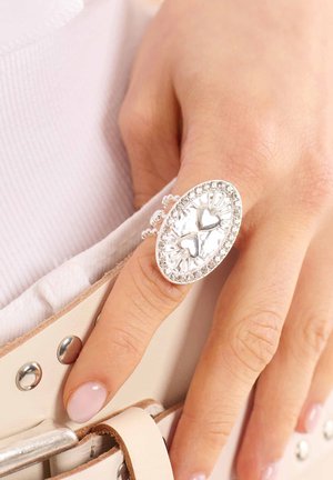 Bague en argent avec un design ovale, featuring deux cœurs au centre et un bord en rhinestones clair. Exposée sur une main près d'une ceinture.