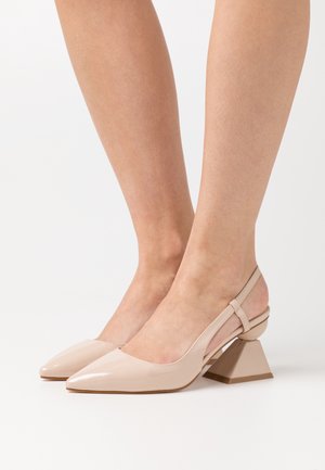 Vrouw draagt naaktkleurige slingback-hooggehakte schoenen van lakleer met puntige neuzen en geometrische blokhakken.