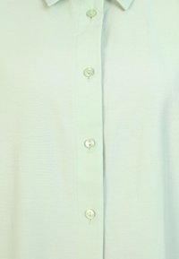 Camisa de botones verde claro con un cuello clásico y seis botones en la parte delantera, con una textura suave y agradable.