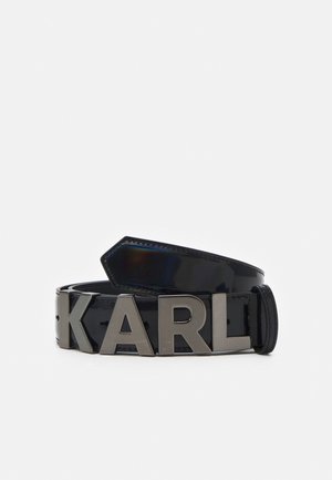 Zwart lakleren riem opgerold met grote metalen letters die KARL spellen op een effen lichte achtergrond.