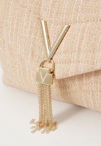 Guldkedja med fransdetalj på en texturerad beige clutchtk bag med en framträdande guldig "V"-logga på ett låsdetalj. Materialet har en subtil lyster.