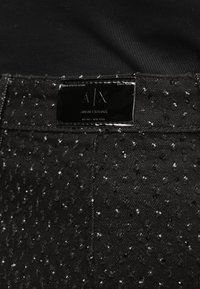 Černá džínová látka s texturovaným povrchem, na kterém jsou malé vystouplé tečky. Na pasu je připevněn lesklý černý štítek s nápisem "A/X" a "Armani Exchange".