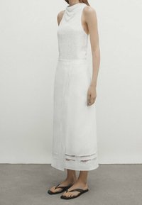 Bezrukawy biały top z drapowanym dekoltem, połączony z długą białą spódnicą z koronkowymi akcentami. Noszony z czarnymi klapkami.