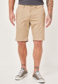 Beige cotton shorts with flat front, side pockets, and stitched hem, paired with gray canvas sneakers and visible ankle tattoos.