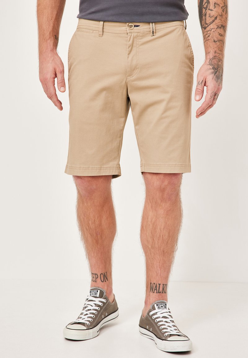 Beige cotton shorts with flat front, side pockets, and stitched hem, paired with gray canvas sneakers and visible ankle tattoos.