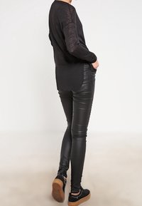 Schwarzer Langarmpullover mit abgerundetem Saum, kombiniert mit engen schwarzen Lederhosen. Das Outfit wird mit schwarzen Sportschuhen mit Nietenakzenten abgerundet.