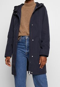 Mörkblå vattentät långrock med huva, tryckknappar och sidofickor, kombinerad med en brun polotröja och blå denimjeans.