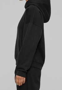 Persona vestida con una sudadera negra y pantalones negros a juego, de pie de lado contra un fondo gris liso.