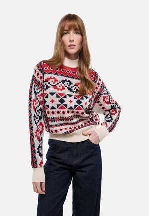 Femme aux longs cheveux bruns portant un pull à motifs géométriques rouges, noirs et blancs et un pantalon taille haute bleu foncé, posant avec une main dans la poche.