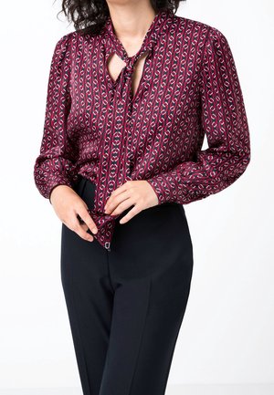 Vrouw draagt een blouse met lange mouwen en een patroon, met een gestrikte halslijn en zwarte getailleerde broek, ze past de zoom van de blouse aan.